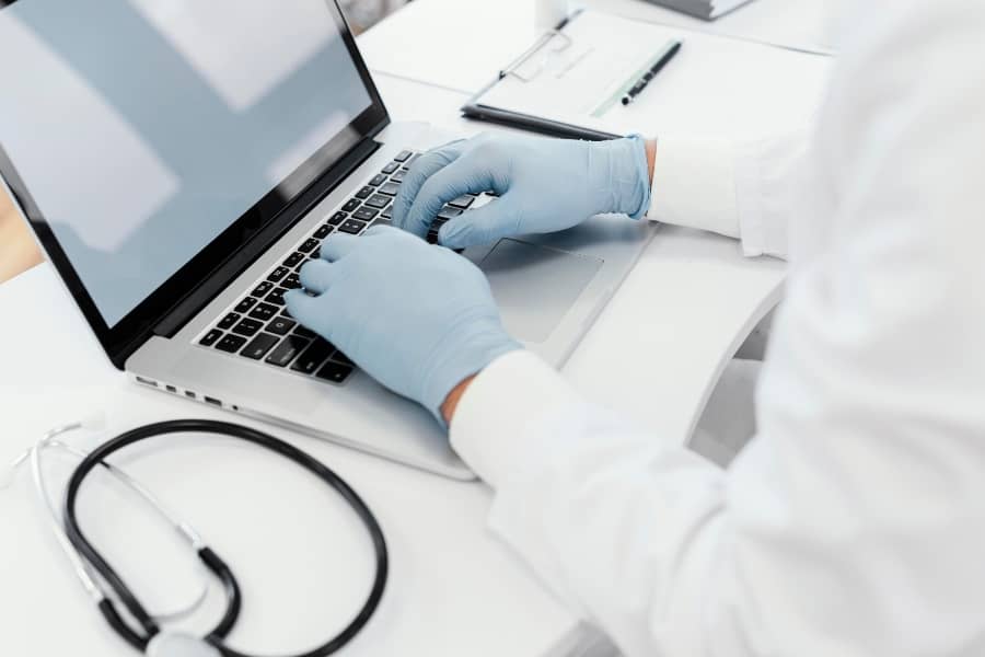 A doctor in a white coat and blue gloves is focused on typing on a laptop to record patient information.